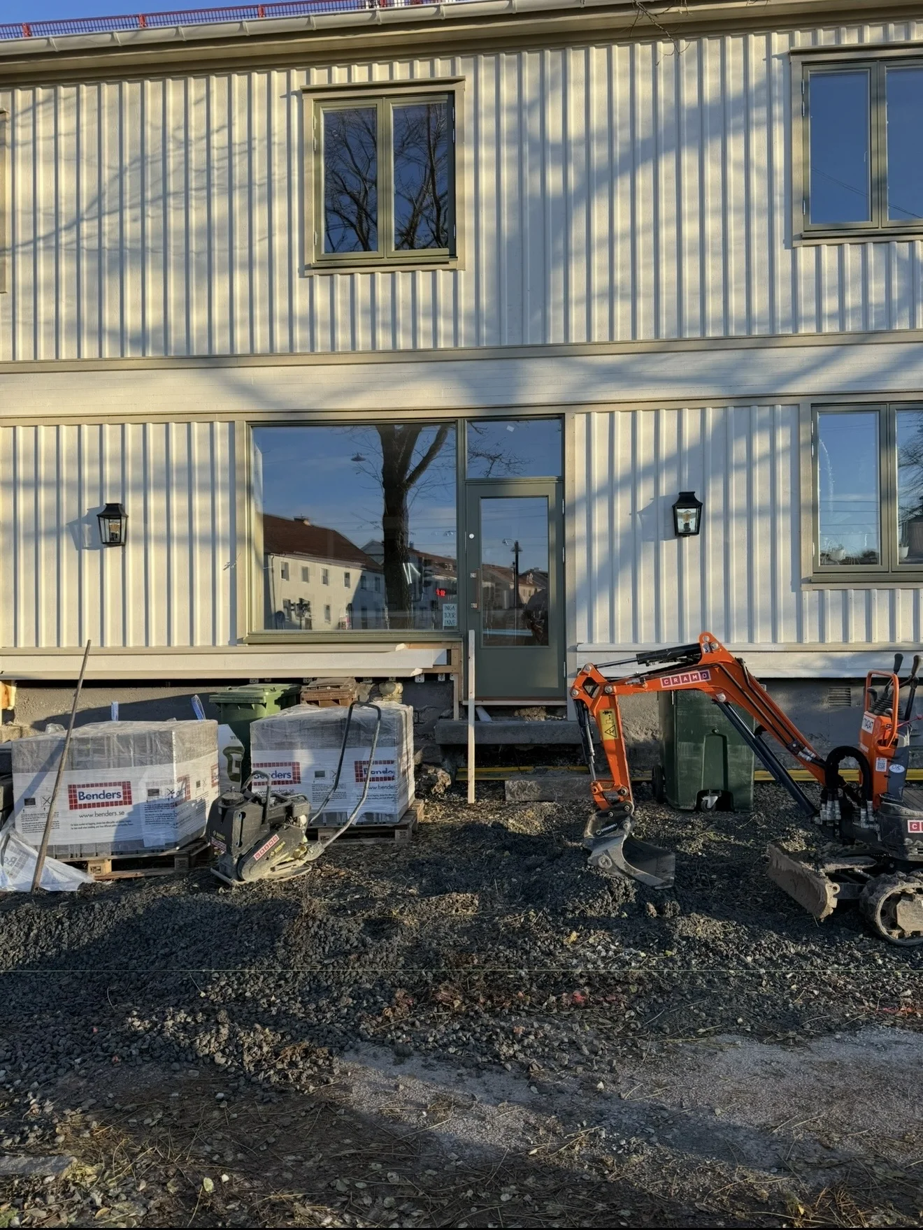 Minigrävare och pallar med marksten framför ett ljust trähus under arbetet