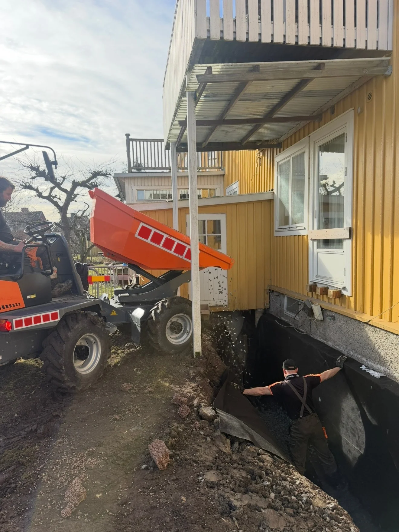Medarbetare monterar fuktskydd på husgrunden medan en dumper tippar massor vid ett gult hus
