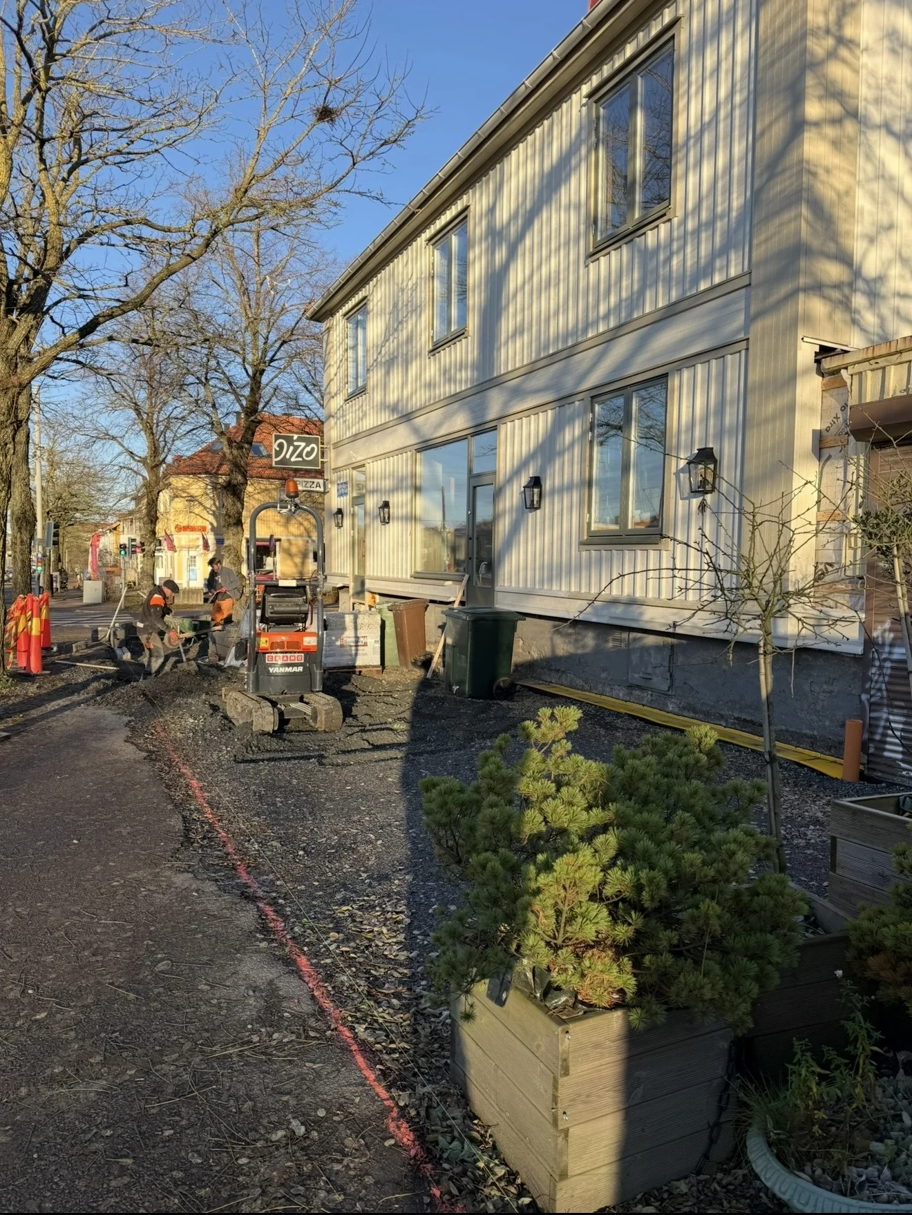 Pågående markarbete längs ett ljust trähus med minigrävare på gatan