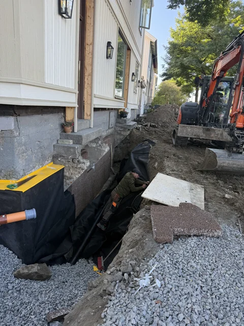Öppet schakt med fuktskyddsmatta längs husgrunden och grävmaskin i bakgrunden